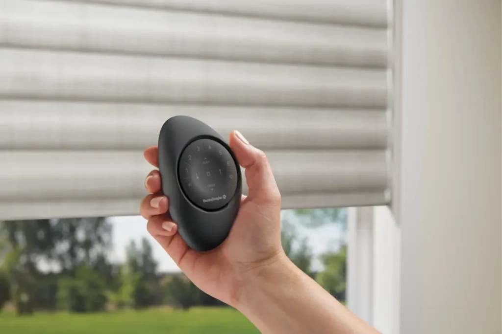 A hand holding the PowerView® Automation Pebble® Remote in a sleek black color by a window